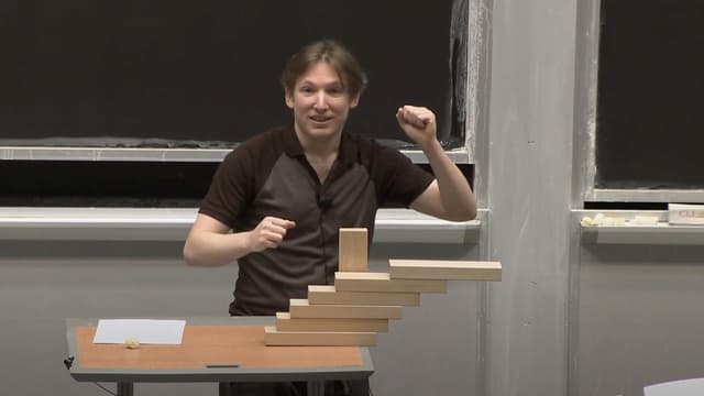 A man stands behind a stack of blocks, each protruding further to the right, with the uppermost block no longer vertically in line with the table in any way.