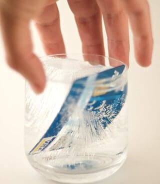 A hand holds a clear glass that contains a credit card frozen in ice.