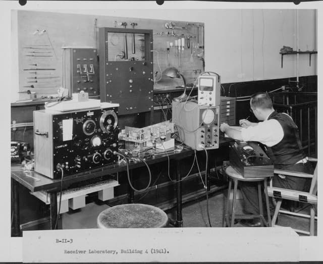 A man writes at a desk which has various large pieces of radio equipment.
